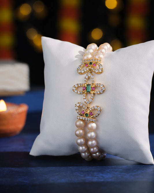 The Bewitching Butterflies Pearl Bracelet by Chandrani Pearls is elegantly showcased on a white cushion, set against a backdrop of softly blurred lights.