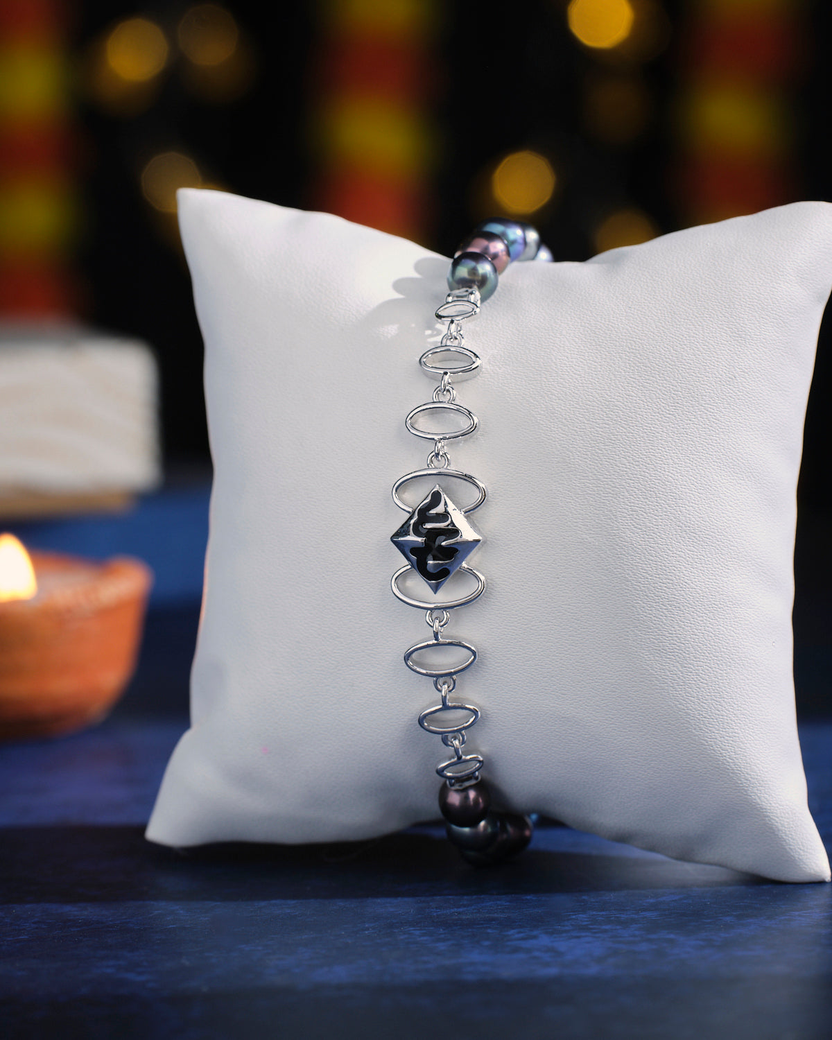 The Curb Link Station Pearl Bracelet by Chandrani Pearls, featuring a silver band and a black centerpiece, is elegantly showcased on a white cushion against a softly blurred dark backdrop.