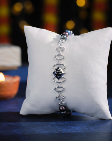 The Curb Link Station Pearl Bracelet by Chandrani Pearls, featuring a silver band and a black centerpiece, is elegantly showcased on a white cushion against a softly blurred dark backdrop.