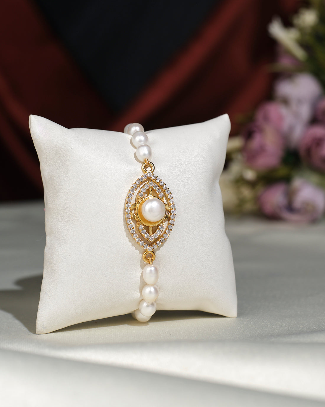 The Trendy Metallic CZ Pretty Bracelet by Chandrani Pearls India features a sparkling centerpiece and is beautifully displayed on a white cushion with blurred flowers in the background—an elegant accessory for any occasion.
