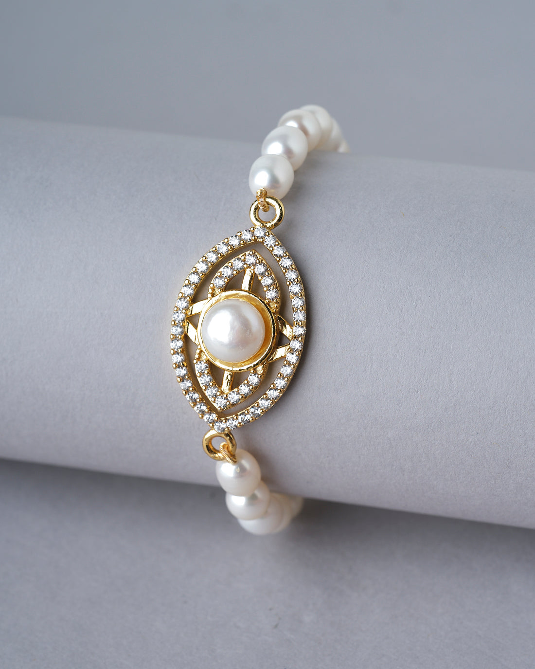 The Trendy Metallic CZ Pretty Bracelet by Chandrani Pearls India features a sparkling centerpiece and is beautifully displayed on a white cushion with blurred flowers in the background—an elegant accessory for any occasion.