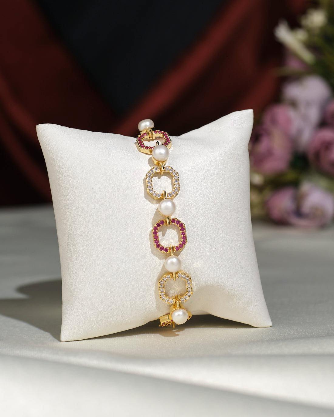 The Fashionable Metallic CZ Pretty Bracelet by Chandrani Pearls India, featuring hexagonal links, small pearls, and red gemstone accents, is displayed elegantly on a white cushion with blurred flowers in the background.