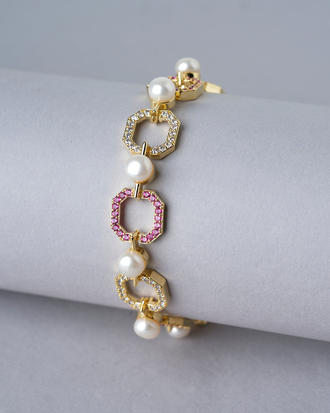 The Fashionable Metallic CZ Pretty Bracelet by Chandrani Pearls India, featuring hexagonal links, small pearls, and red gemstone accents, is displayed elegantly on a white cushion with blurred flowers in the background.