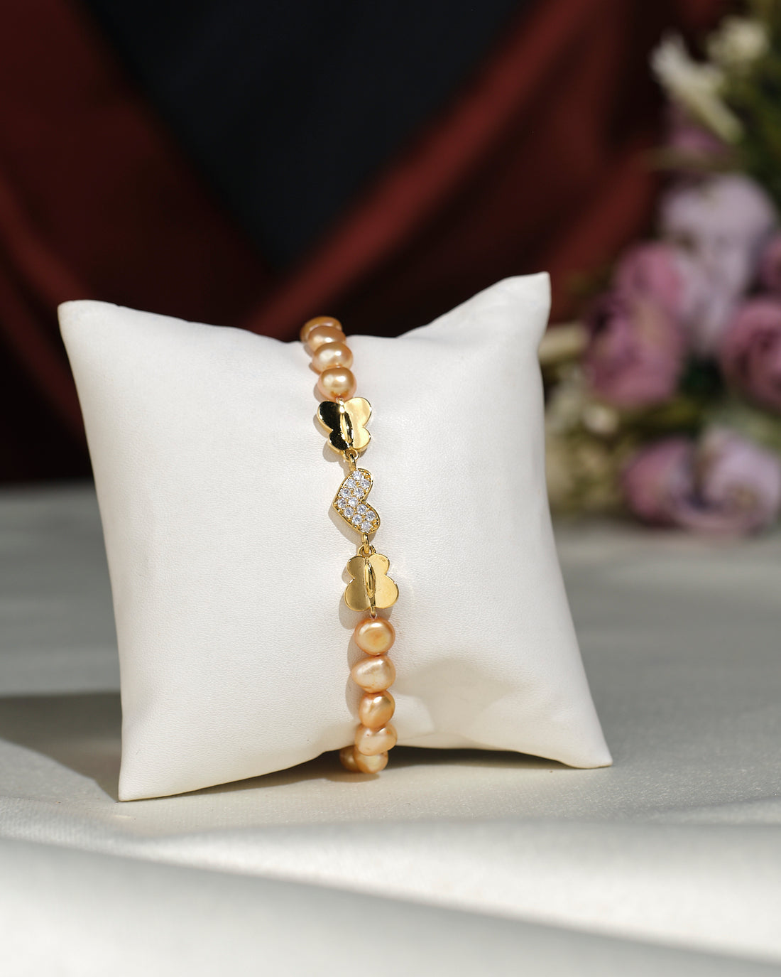 Little Heart CZ Pearl Bracelet by Chandrani Pearls India shown on a white cushion, featuring pearls and a decorative clasp, with blurred flowers in the background.