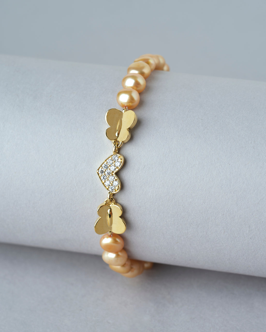 Little Heart CZ Pearl Bracelet by Chandrani Pearls India shown on a white cushion, featuring pearls and a decorative clasp, with blurred flowers in the background.