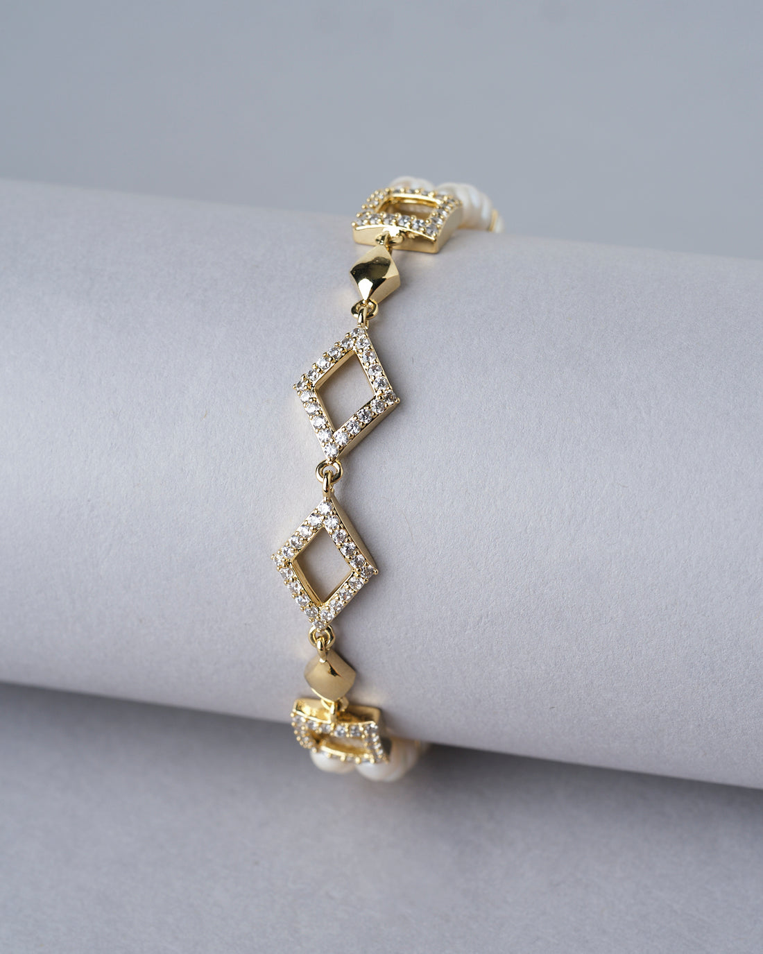 The Regal Pearl Bracelet by Chandrani Pearls India showcases elegant gold and diamond geometric design with pearl accents, displayed on a white cushion with soft, blurred flowers in the background.
