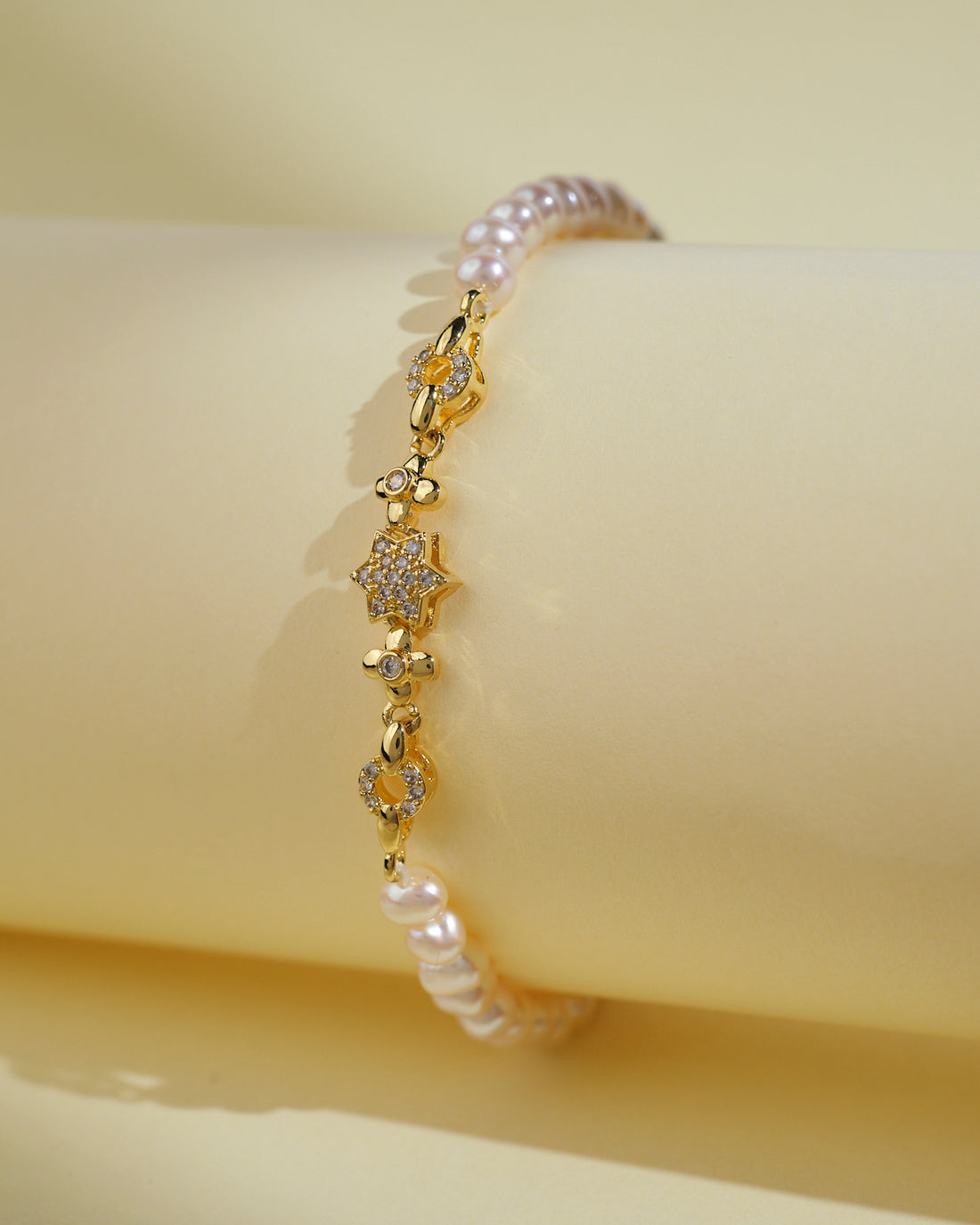 The Flory White 1 Line Pearl Bracelet by Chandrani Pearls India features gold, small floral designs, and clear stones for everyday elegance. Shown against a pale yellow background.