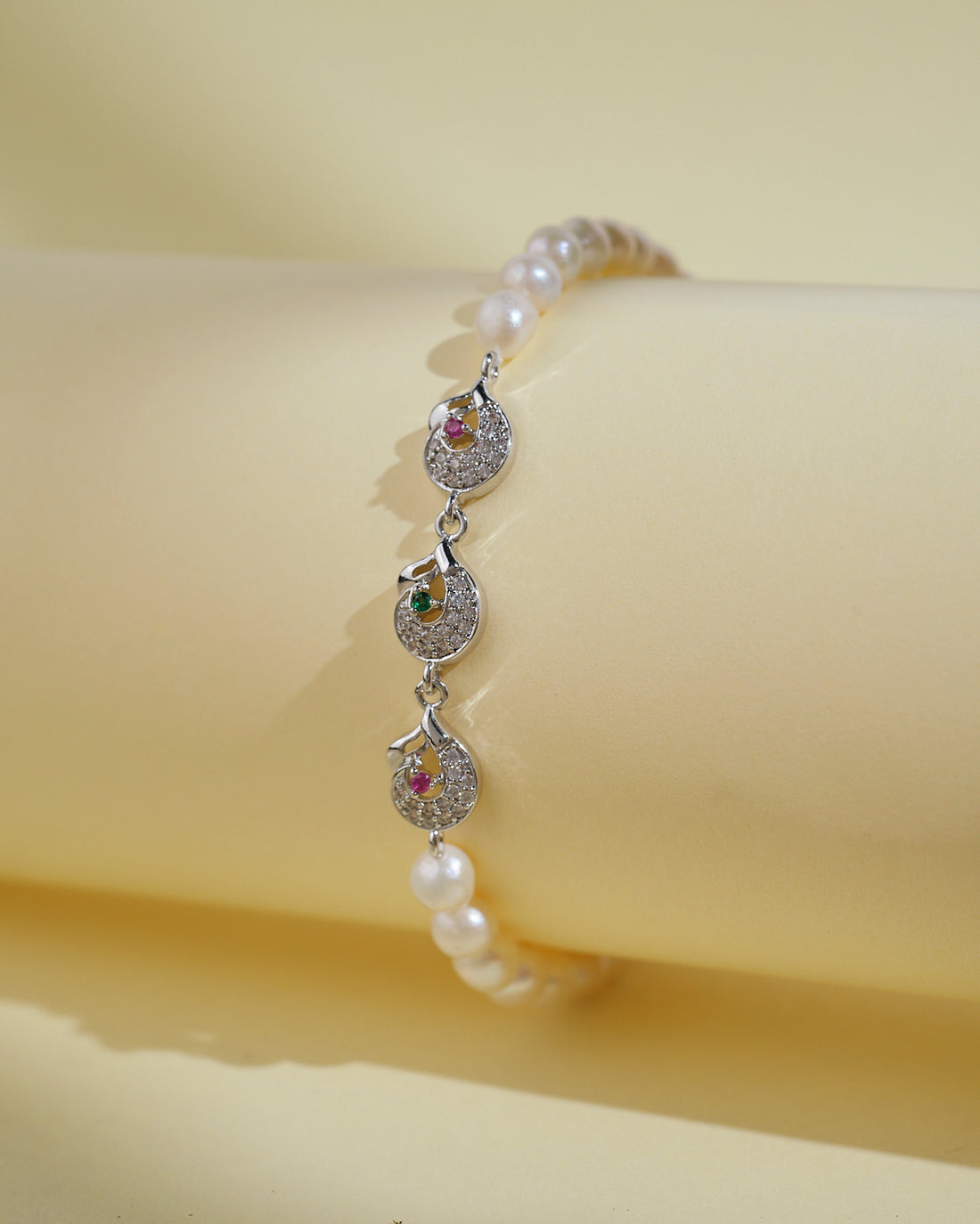 The Pretty White 1 Line Pearl Bracelet by Chandrani Pearls India features elegant pearls and three silver pendants with colored stones, shown on a light yellow background—an ideal choice for everyday luxury.