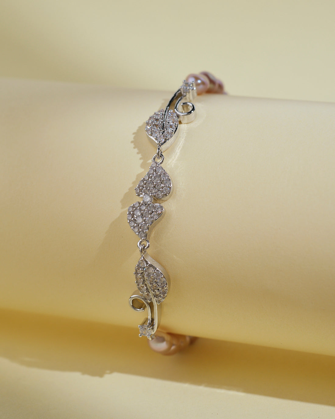 The Leafy Pink1 Line Pearl Bracelet by Chandrani Pearls India, featuring leaf-shaped links and pearls, displayed on a light yellow cylindrical surface, embodies the elegance and simplicity of refined jewelry design.