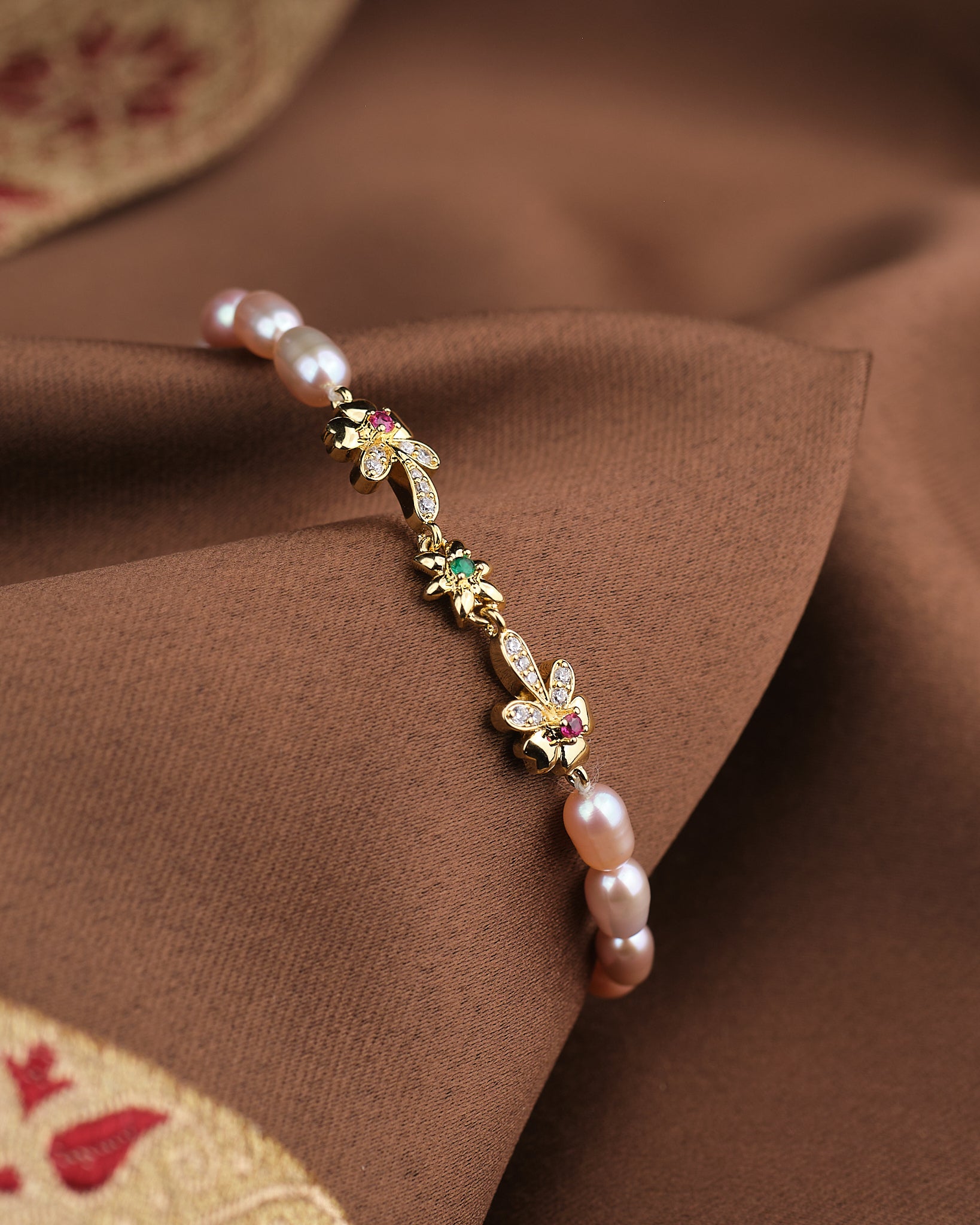 The Noor Classic Pearl Radiance Bracelet by Chandrani Pearls features hand-selected pearls, CZ stones, and gold floral and leaf charms, elegantly displayed on brown fabric with a gold embroidered edge.