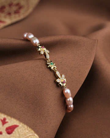 The Noor Classic Pearl Radiance Bracelet by Chandrani Pearls features hand-selected pearls, CZ stones, and gold floral and leaf charms, elegantly displayed on brown fabric with a gold embroidered edge.