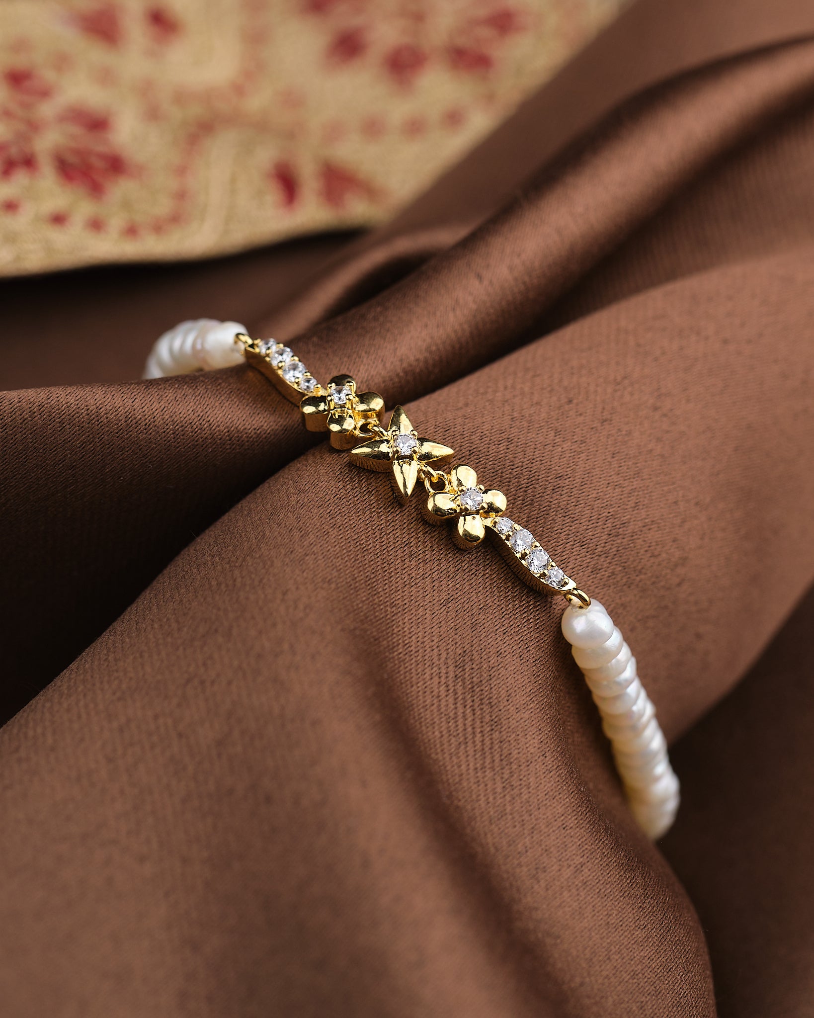 The Noor Elegant Harmony Bracelet by Chandrani Pearls, featuring floral details, delicate pearls, and sparkling CZ stones, is displayed on brown fabric.