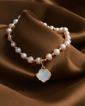 The Classic Elegance bracelet by Chandrani Pearls, featuring beaded pearls and a mother-of-pearl clover pendant with gold accents, adds a timeless shimmer to any style as it rests on brown fabric.