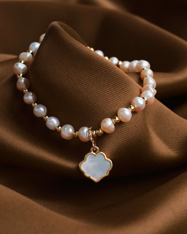 The Classic Elegance bracelet by Chandrani Pearls, featuring beaded pearls and a mother-of-pearl clover pendant with gold accents, adds a timeless shimmer to any style as it rests on brown fabric.
