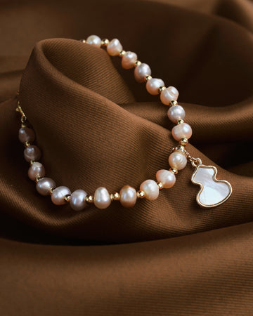 The Elegant Mother of Pearl Charm Bracelet by Chandrani Pearls features gold accents and a white guitar-shaped pendant, displayed on soft brown fabric.