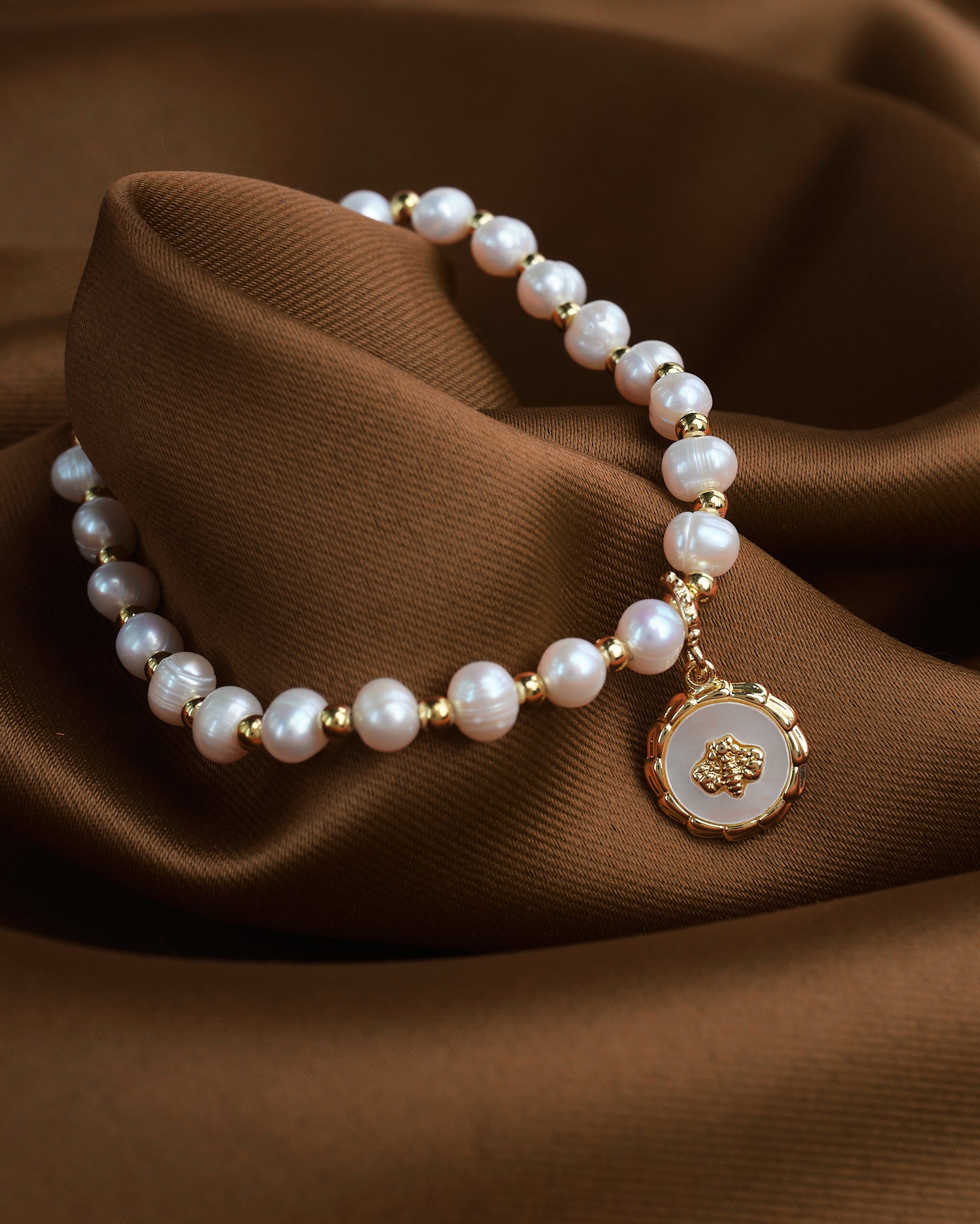 The Beautiful Mother of the Pearl Charm Bracelet by Chandrani Pearls, featuring gold beads and a round pendant, rests elegantly on brown fabric.