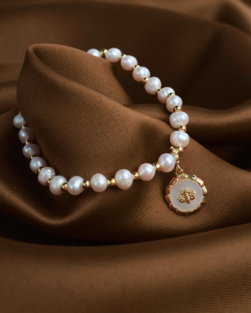 The Beautiful Mother of the Pearl Charm Bracelet by Chandrani Pearls, featuring gold beads and a round pendant, rests elegantly on brown fabric.