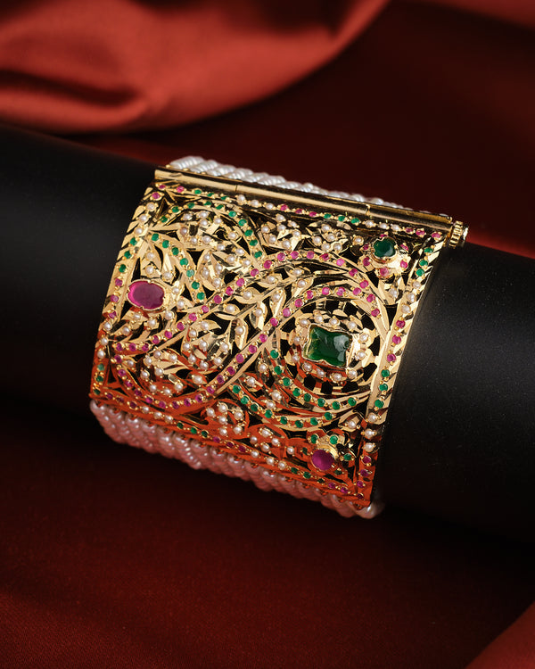 The Scintillating Stone With Style Pearl Bracelet by Chandrani Pearls, featuring green and pink gemstones in a gold filigree design, is showcased on a black stand with a rich red fabric background.
