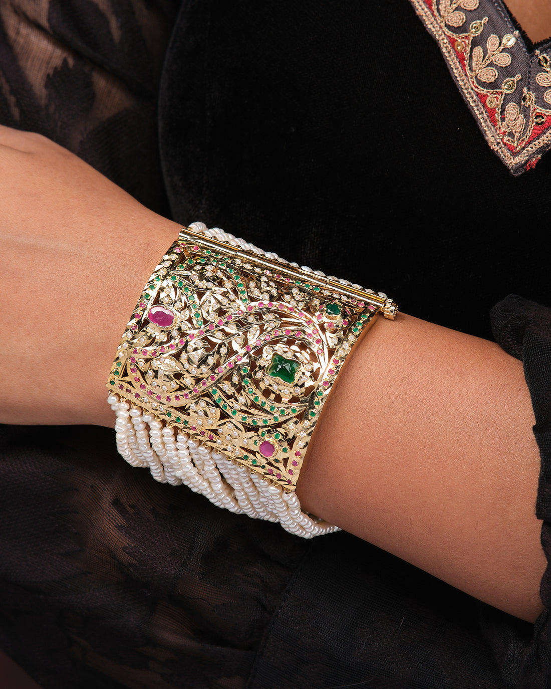 The Scintillating Stone With Style Pearl Bracelet by Chandrani Pearls, featuring green and pink gemstones in a gold filigree design, is showcased on a black stand with a rich red fabric background.