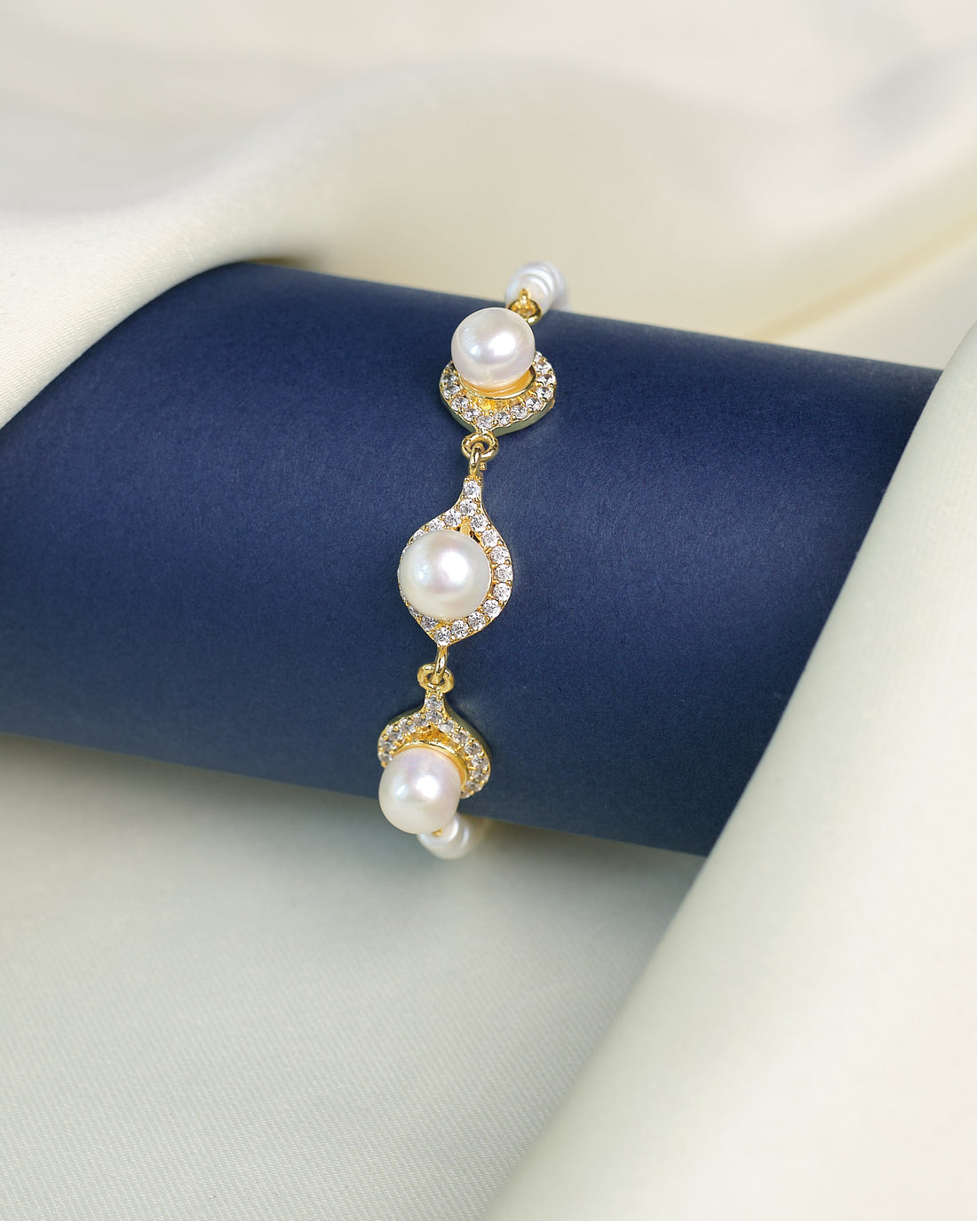 The Classic Beauty Pearl Bracelet with CZ Detailing from Chandrani Pearls is displayed on a dark blue cylindrical stand, surrounded by cream-colored fabric. Elegant pearls and sparkling cubic zirconia create a timeless look.