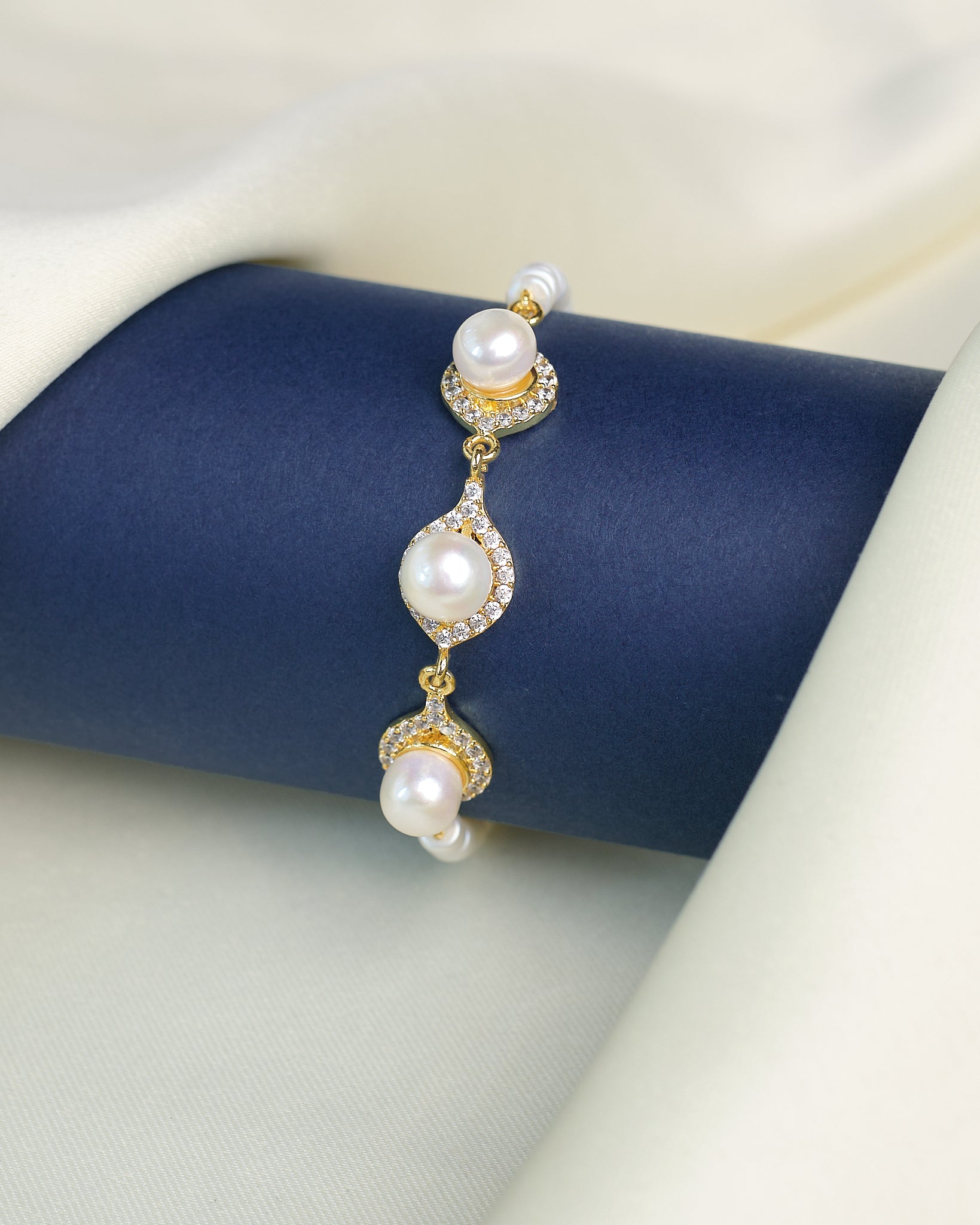 The Classic Beauty Pearl Bracelet with CZ Detailing from Chandrani Pearls is displayed on a dark blue cylindrical stand, surrounded by cream-colored fabric. Elegant pearls and sparkling cubic zirconia create a timeless look.