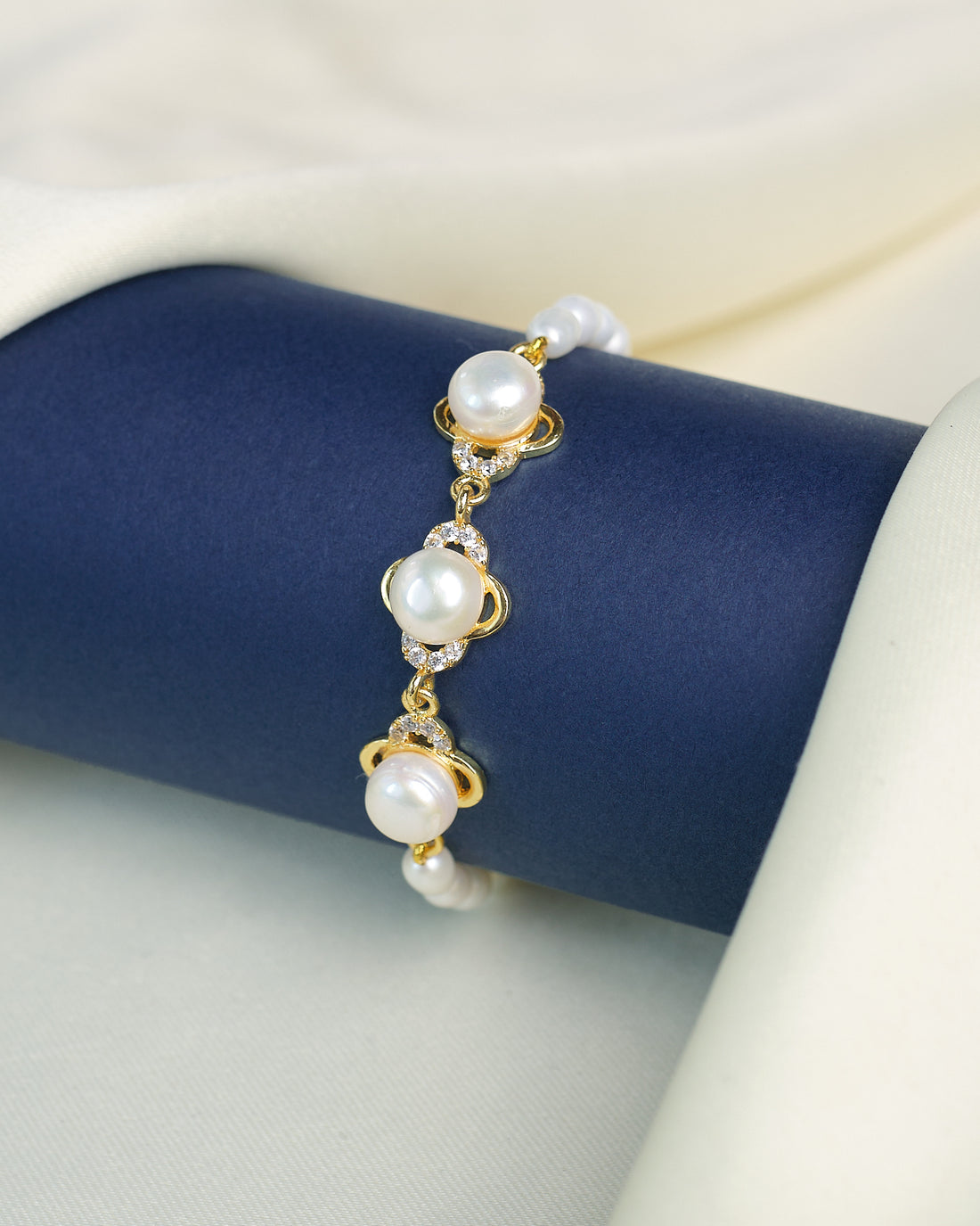 The Chandrani Pearls Refined Pearl Bracelet with Brilliant CZ Stones showcases round white pearls and sparkling cubic zirconia, elegantly displayed on a navy cylinder against a cream fabric background.