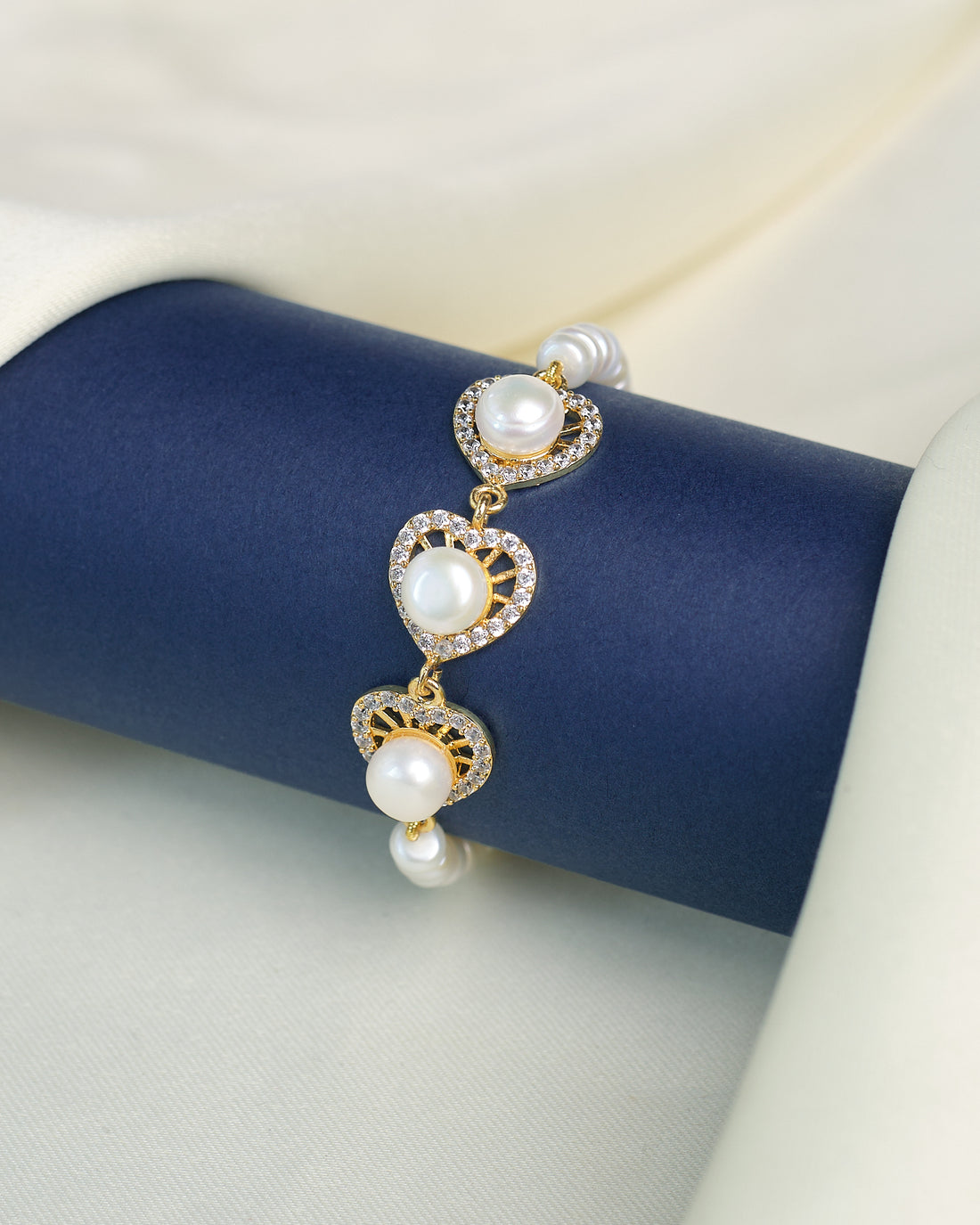 The Modern Pearl Bracelet with Cubic Zirconia Highlights from Chandrani Pearls features three gold, heart-shaped links adorned with pearls and sparkling stones—an elegant choice for wedding jewelry, shown on a dark blue display against light fabric.