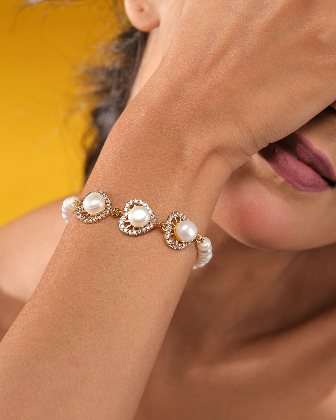 The Modern Pearl Bracelet with Cubic Zirconia Highlights from Chandrani Pearls features three gold, heart-shaped links adorned with pearls and sparkling stones—an elegant choice for wedding jewelry, shown on a dark blue display against light fabric.