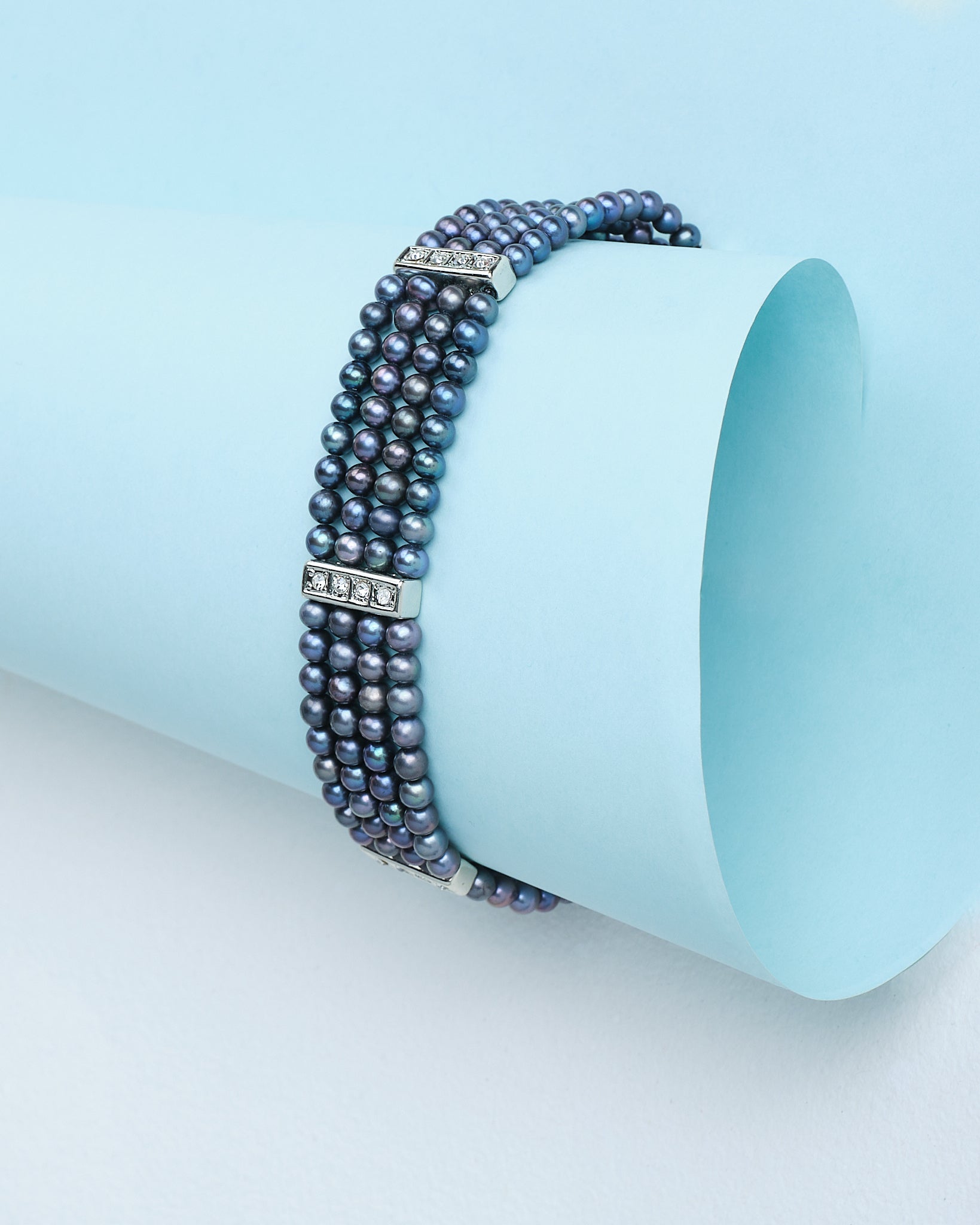 The "A Touch of Elegance on Your Wrist" bracelet by Chandrani Pearls features three rows of sophisticated pearls with rhinestone-studded spacers, displayed on a rolled sheet of light blue paper.