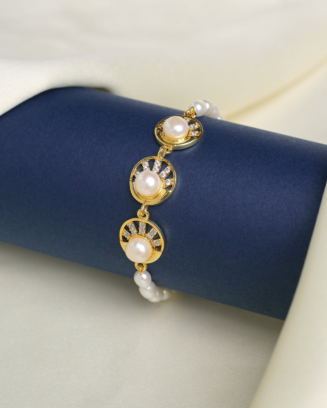 The Chandrani Pearls Radiant Pearl Bracelet with High-Polish CZ features three round pearls and sparkling cubic zirconia, shown on a dark blue cylindrical stand against cream fabric to showcase its refined finish.