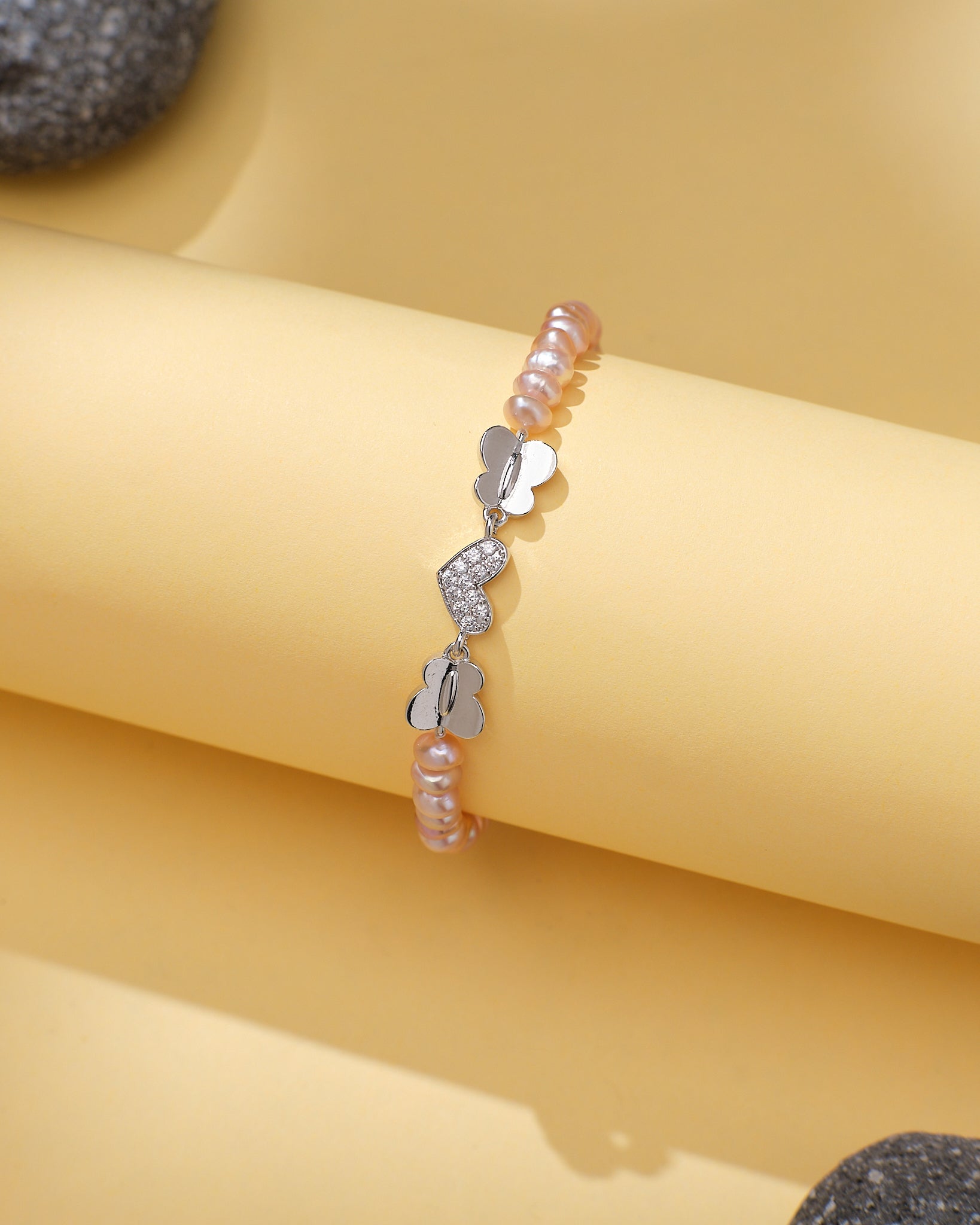 The Chandrani Pearls Elegant Pearl & CZ Tennis Bracelet Pink Pearl features pink pearls, silver butterfly charms, and sparkling cubic zirconia, displayed on a curved yellow surface with gray stones nearby.