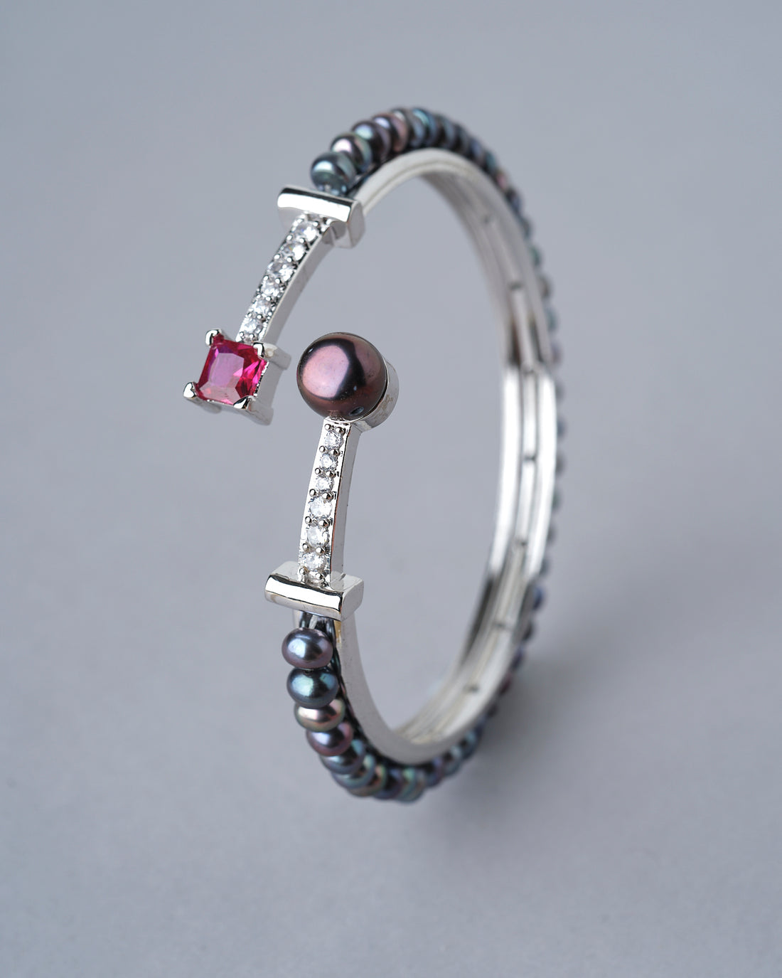 The Trendy Black Pearl Bangle by Chandrani Pearls India, featuring a silver band with a red square gemstone, round glowing pearl, black beads, and clear stones, is displayed upright on a white circular stand.