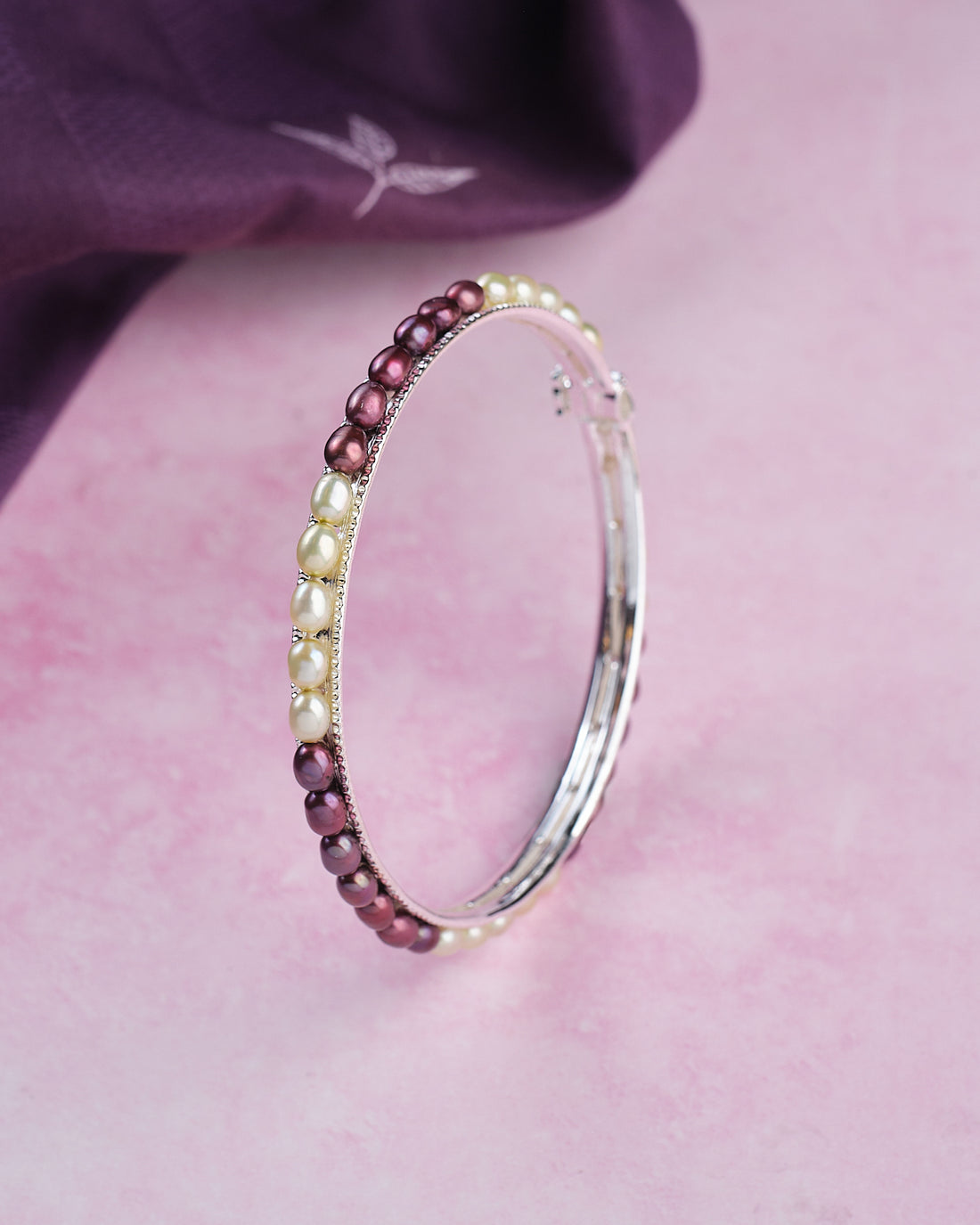The Chandrani Pearls Single Line Black & White Bangle (B01731) stands upright on a pink surface, with a purple cloth in the background.