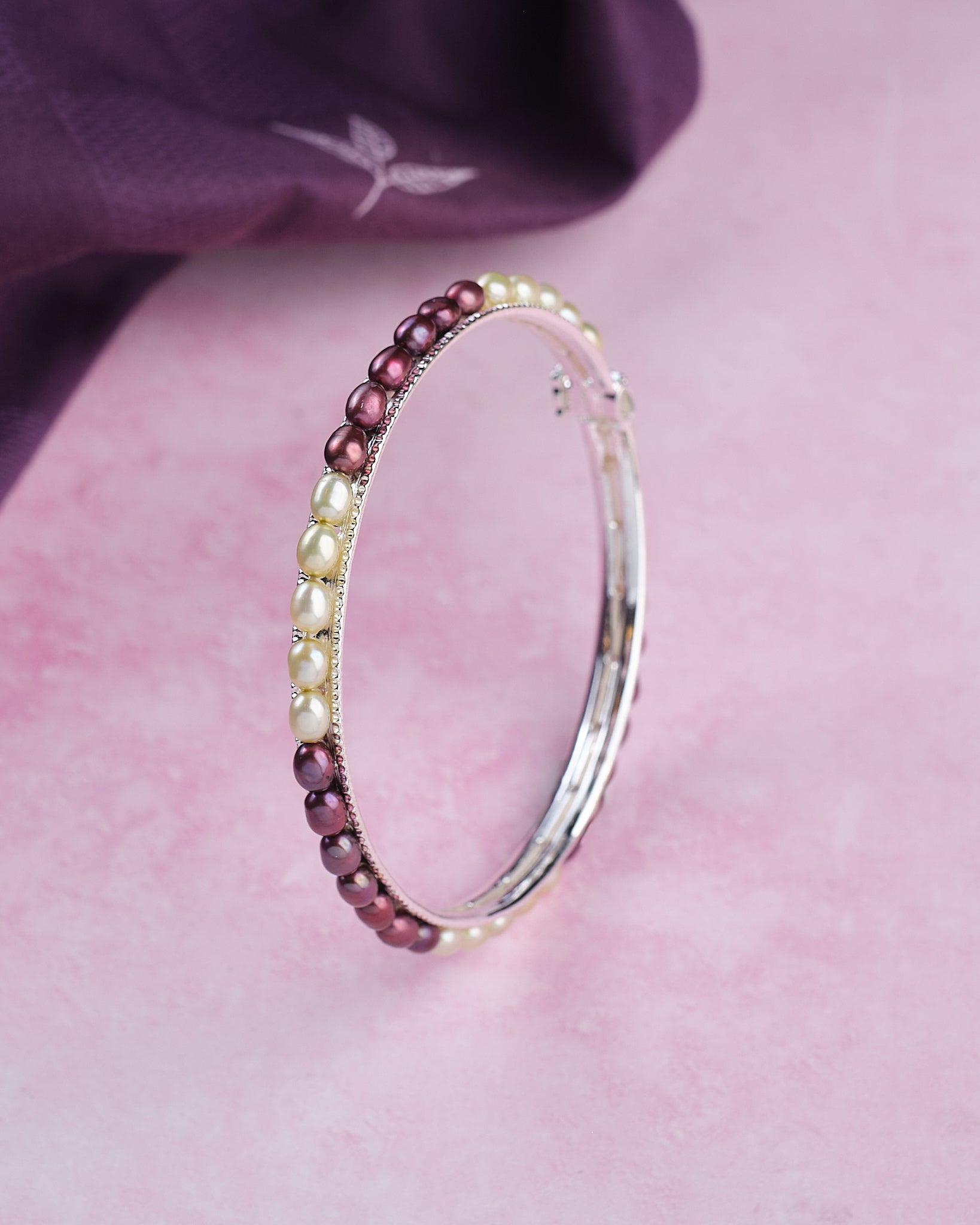 The Chandrani Pearls Single Line Black & White Bangle (B01731) stands upright on a pink surface, with a purple cloth in the background.