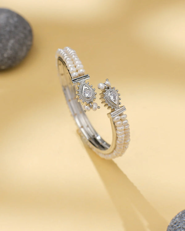 The Chandrani Pearls Silver-Plated Pearl and CZ Designer Pearl Bangle, featuring small pearls and teardrop CZ accents, is displayed on a light beige surface with a smooth stone nearby.