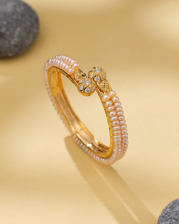 The Classic Pearl Bangle with CZ Accents Bridal Perfection by Chandrani Pearls, featuring delicate pearls and CZ-studded ends, is displayed on a beige surface with smooth gray stones as stylish accents.