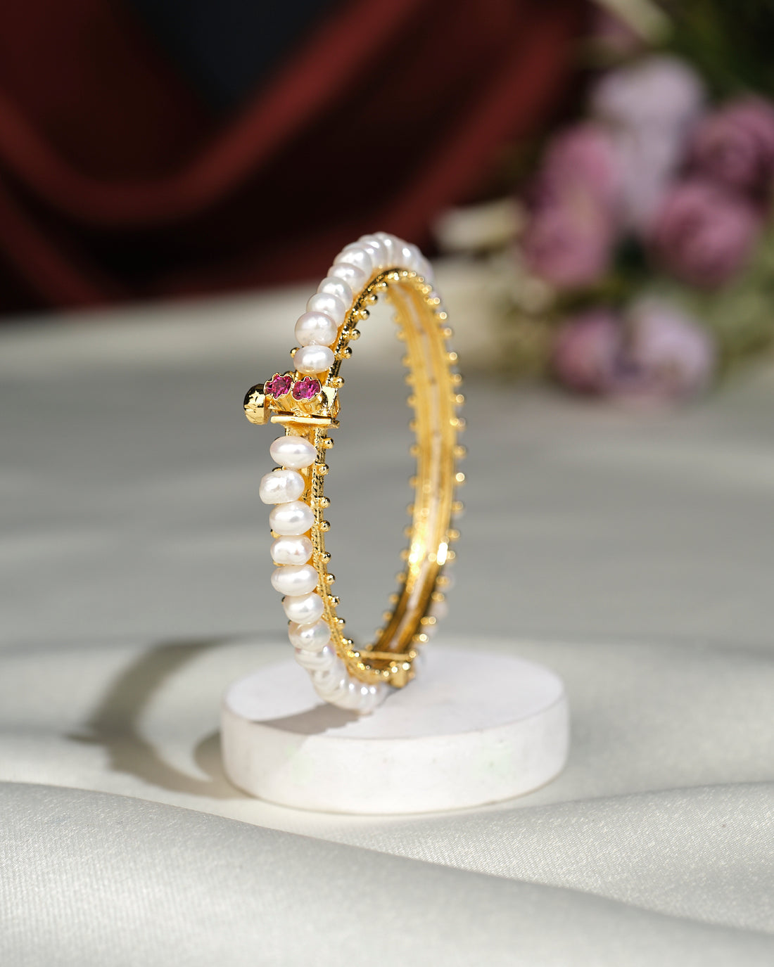 The Simple Pearl Bangle by Chandrani Pearls India, featuring small pearls and a central pink gemstone, is displayed upright on a round white stand against a blurred floral background.