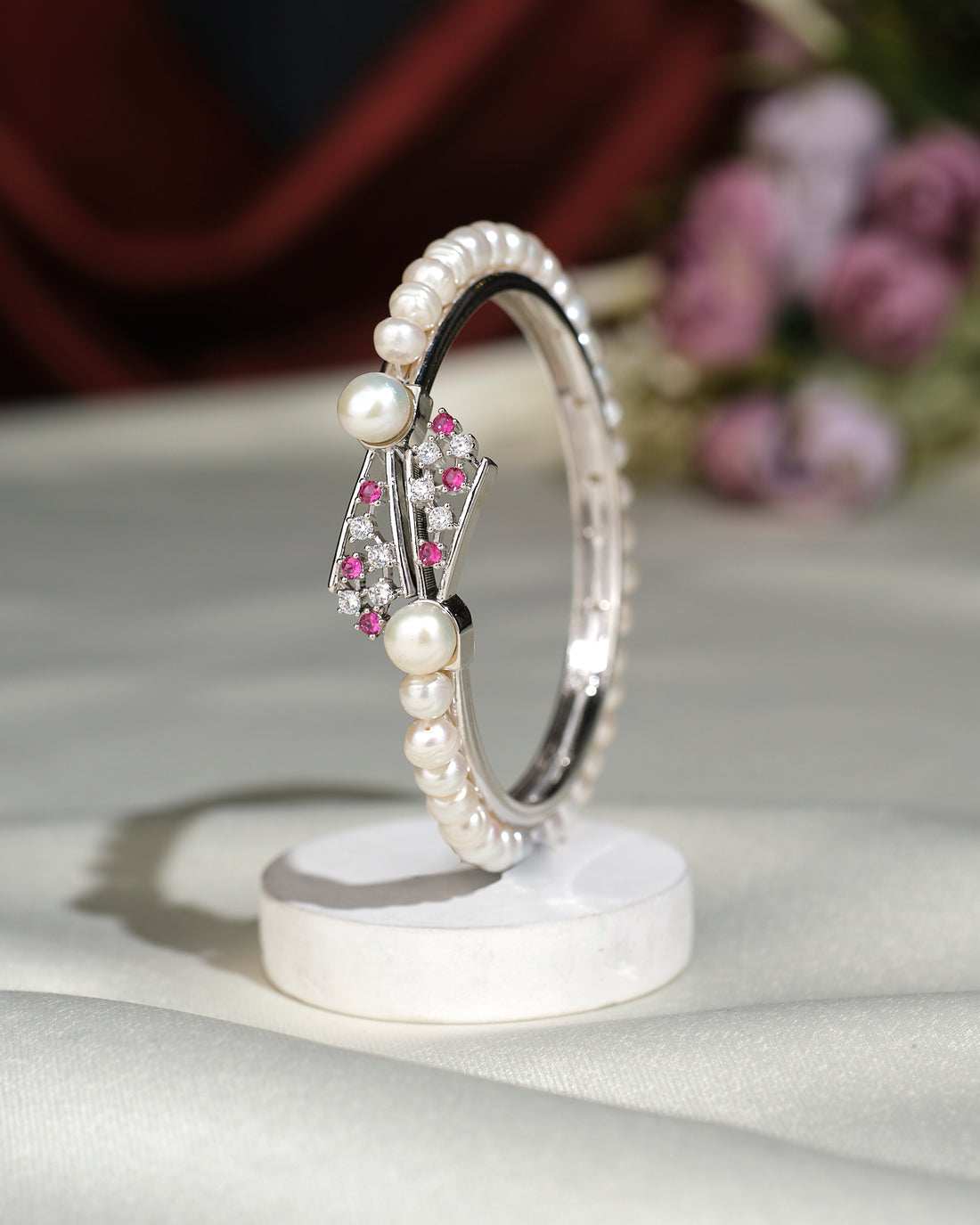 The Trendy White Pearl Bangle by Chandrani Pearls India features a row of white pearls with pink gemstone accents and two larger pearls, shown upright on a white circular stand against a blurred floral backdrop.