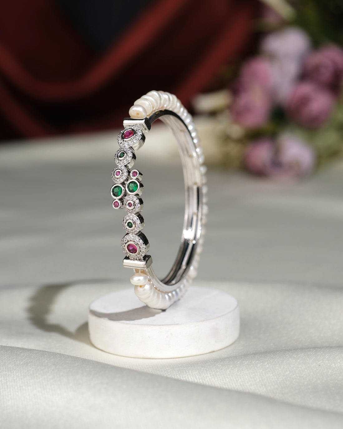 The Fancy White Pearl Bangle by Chandrani Pearls India, featuring pearl accents and multicolored gemstones, is displayed upright on a round white stand with blurred flowers in the background.