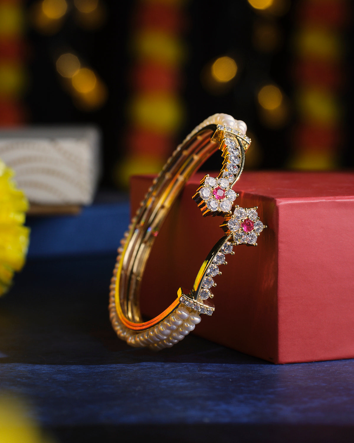 The Ragapushpi - The Rosy Night Pearl Bangle by Chandrani Pearls, adorned with pearl accents and floral embellishments, featuring pink and clear stones, is elegantly displayed on a red box against a blurred background.