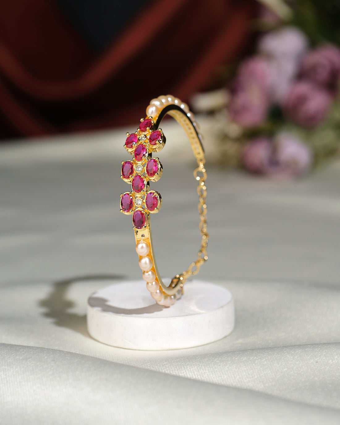 The Pretty Pink Bangle by Chandrani Pearls India, featuring pink gemstones and white pearls set in elegant gold, stands upright on a round white base against a blurred floral background.