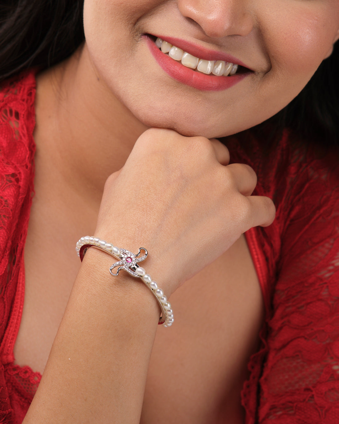 The Trendy White Bangle by Chandrani Pearls India features an elegant silver design with a pink gemstone centerpiece, displayed on a white stand with blurred flowers in the background.