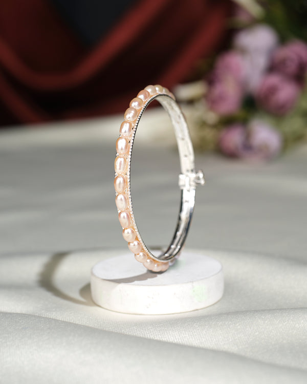 The Simple Pearl Bangle by Chandrani Pearls India, featuring a sleek silver design with small pearls, is elegantly displayed upright on a round white stand against a softly blurred background with purple flowers.
