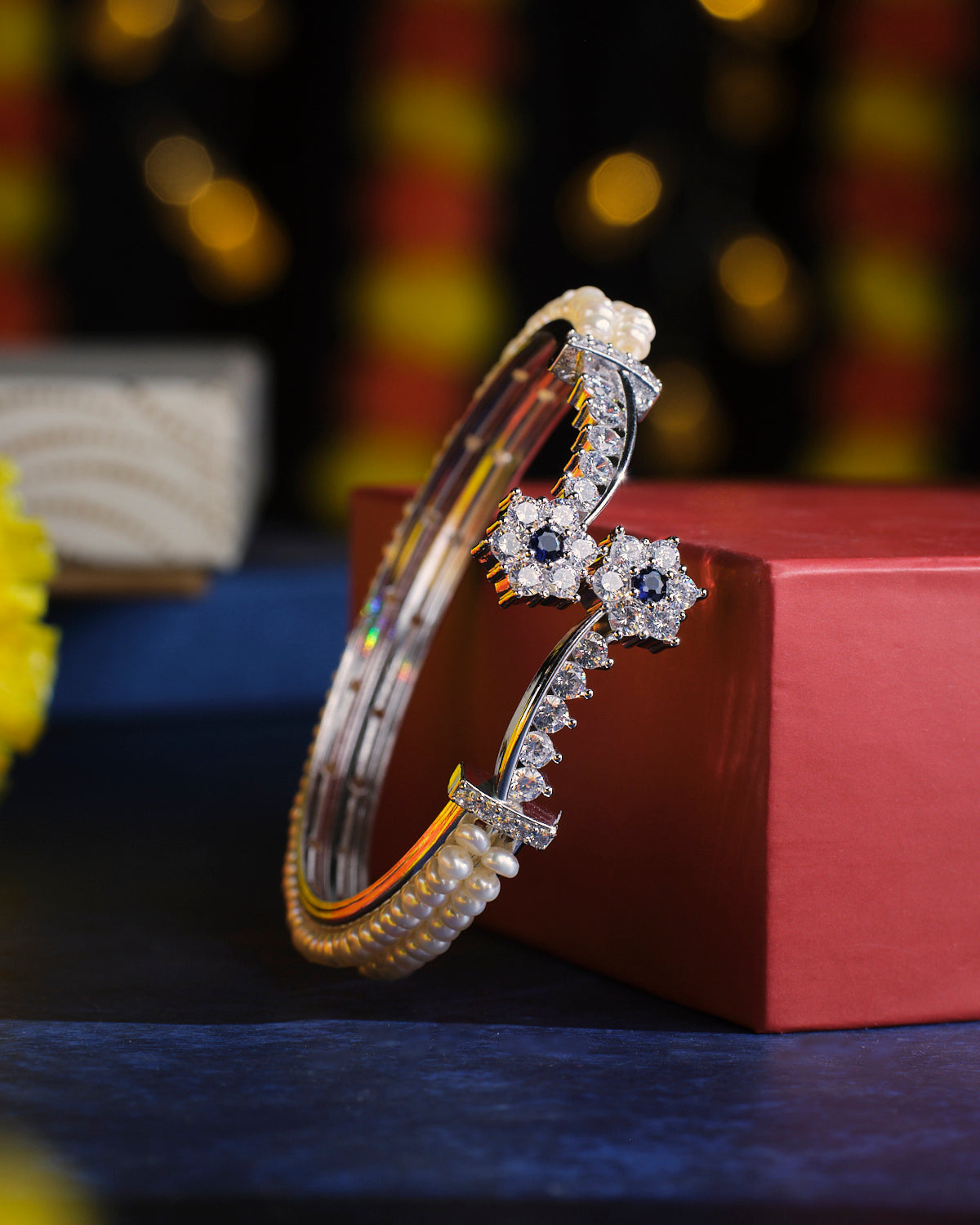 The Buzzing Sweetness Pearl Bangle from Chandrani Pearls, embellished with pearls and star-shaped accents, is elegantly displayed on a red box against a bokeh background.