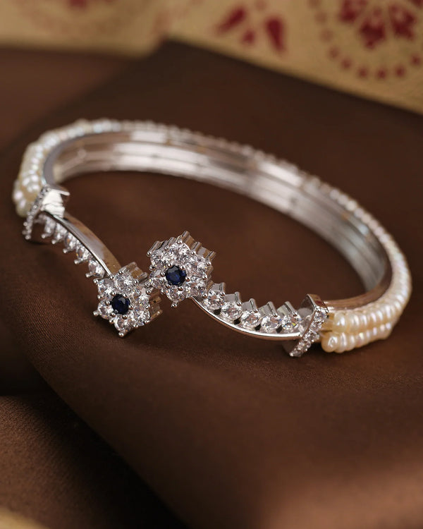 The Chandrani Pearls Floral Blossom Pearl CZ Bangle features small pearls, two star-shaped accents with blue and clear stones, and delicate floral CZ motifs, elegantly displayed on brown fabric.