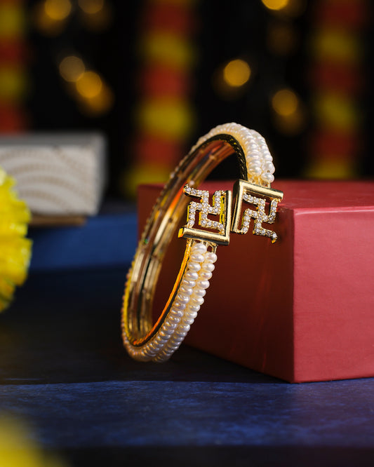 The Shubh Swastik Pearl Bangle by Chandrani Pearls, featuring gold accents and decorative elements, is elegantly displayed on a red box against a blurred background.