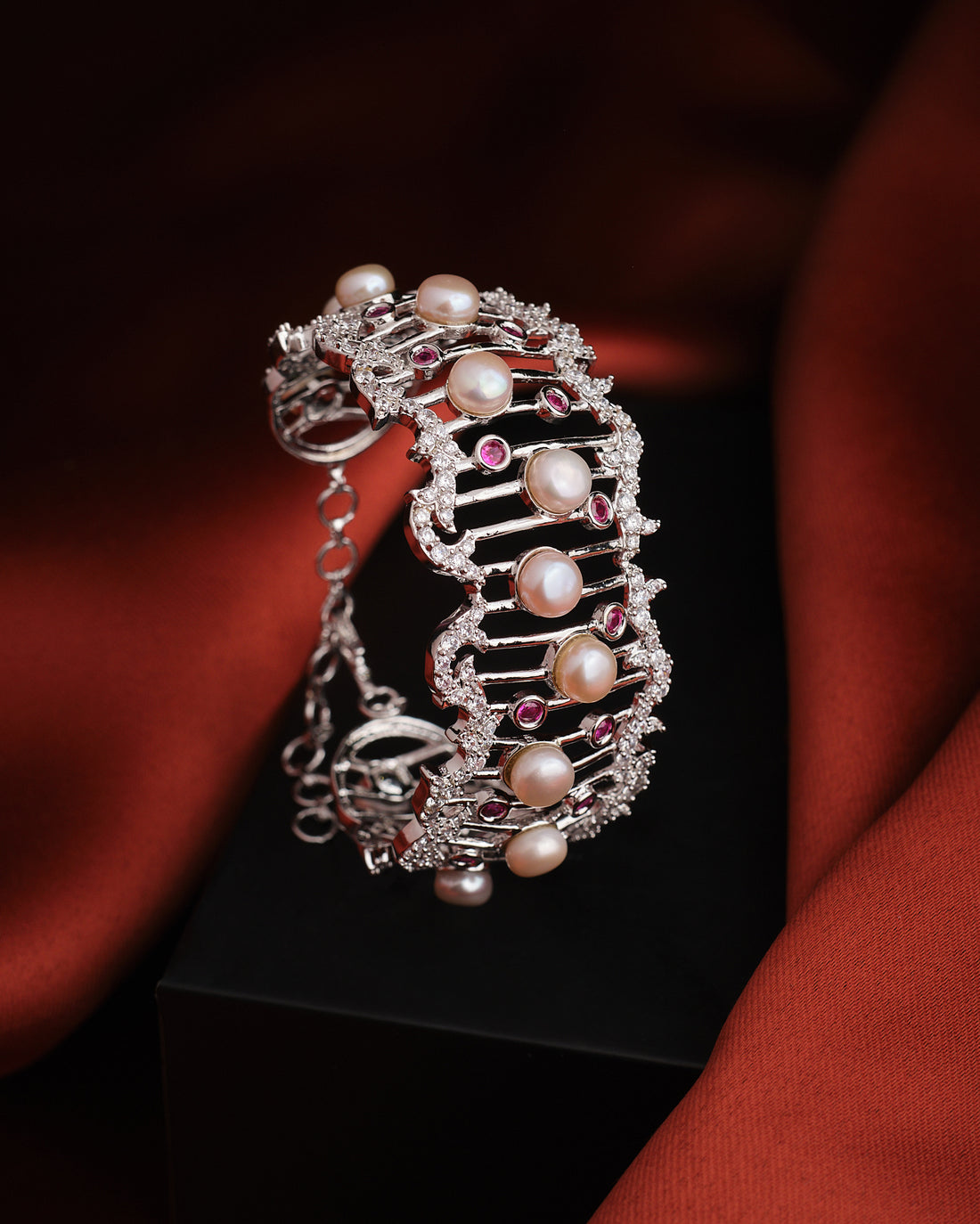 The Beauty of Rezi Pearl Bangle by Chandrani Pearls, featuring white pearls and red gemstones, is elegantly displayed on a black box with a dark red fabric background—an ideal choice for memorable occasions.