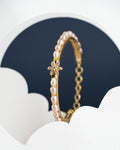 The Astoria Tulip Pearl Bangle by Chandrani Pearls India is a gold bracelet for kids featuring pearls and a floral design, set against a dark backdrop with white cloud-like patterns.