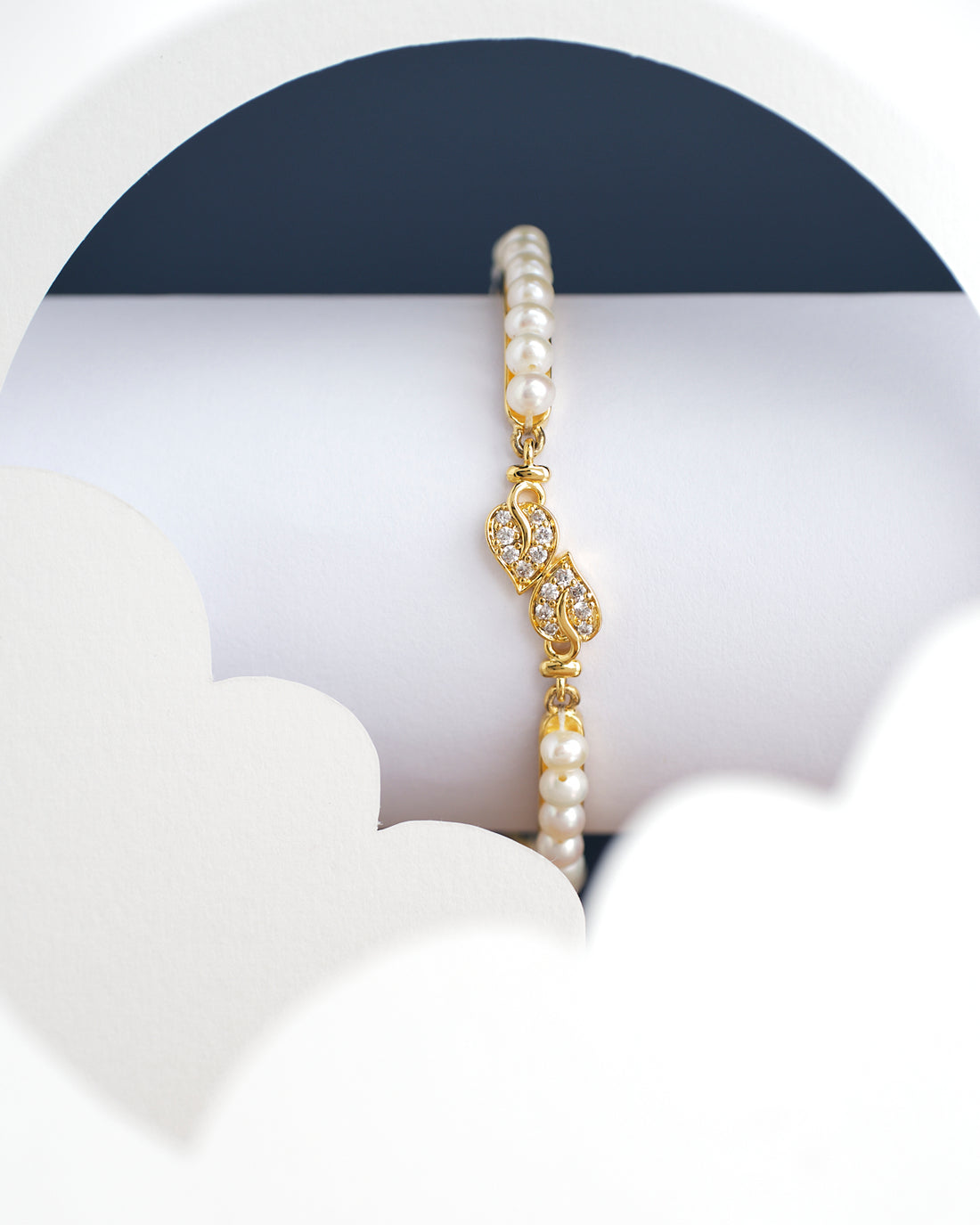 The Leafy Pearl Bangle for kids by Chandrani Pearls India, featuring a gold clasp, is showcased against a white and dark blue abstract background.