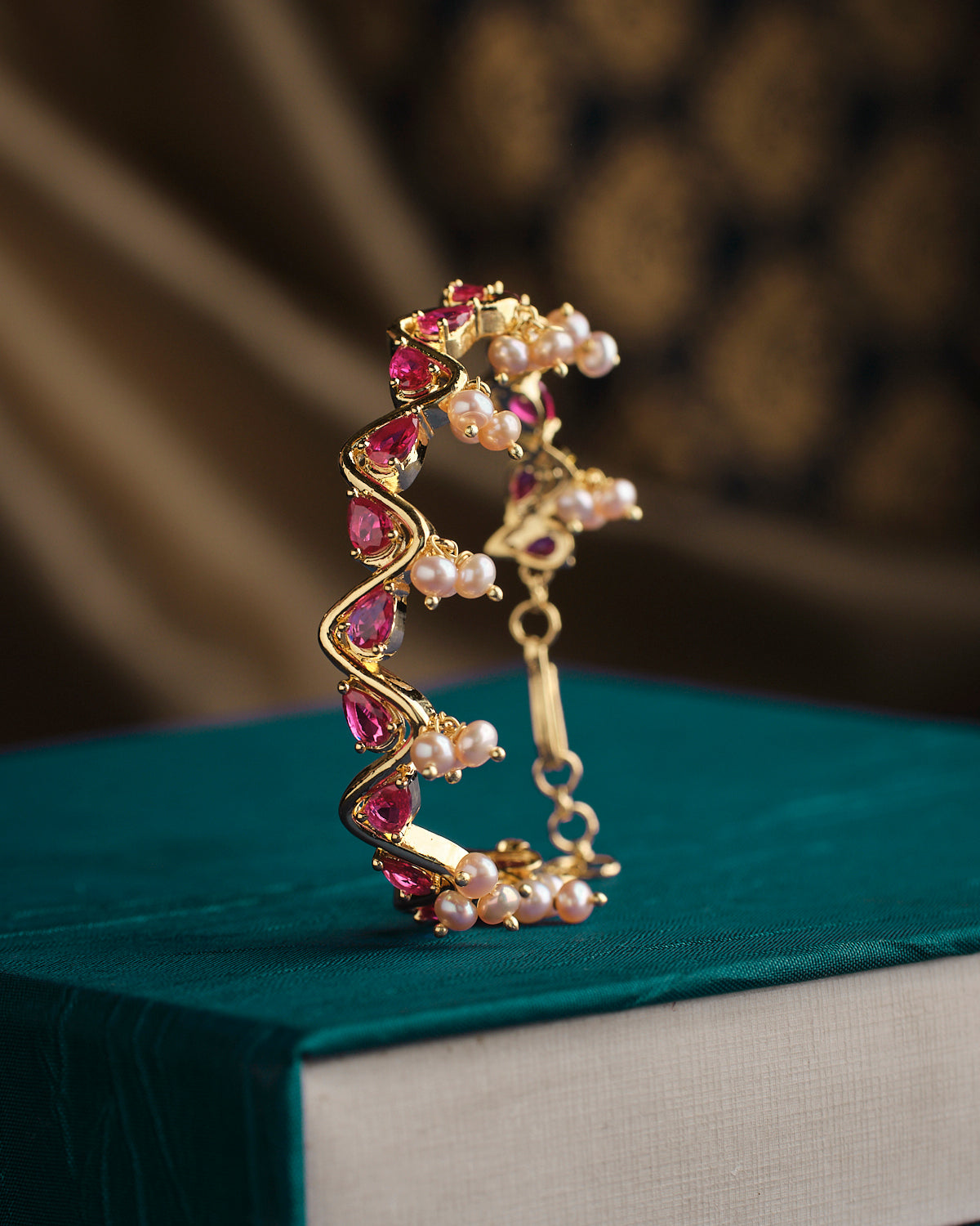 The Unify Toggle Bangle by Chandrani Pearls, adorned with red gemstones and pearls, is showcased on a teal box with an intricate design and a decorative background.